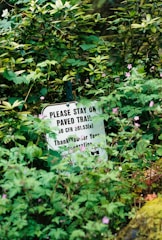 A sign with text partially hidden among dense vegetation. The visible text advises staying on paved trails and thanks for cooperation, surrounded by various green plants and small pink flowers.