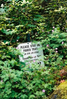 A sign with text partially hidden among dense vegetation. The visible text advises staying on paved trails and thanks for cooperation, surrounded by various green plants and small pink flowers.