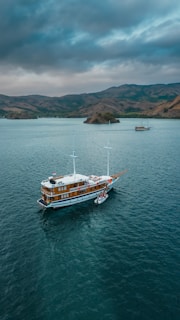 A large wooden sailboat floats in a calm body of water with lush hills in the background. The sky is overcast with dark clouds, adding a sense of impending weather change. A small dinghy is docked alongside the sailboat. Another smaller boat is visible in the distance near the shoreline.