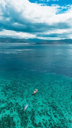 Two small boats are floating on a crystal clear turquoise ocean near the shore. The vibrant coral reefs are visible beneath the surface, while the horizon features a majestic mountain range under a sky filled with expansive clouds.
