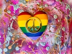 A heart-shaped object painted with the rainbow colors of the LGBTQ+ pride flag, featuring a prominent gold peace symbol in the center. The background is a vibrant and chaotic mix of bright pinks, purples, and oranges splattered with metallic silver and gold textures.