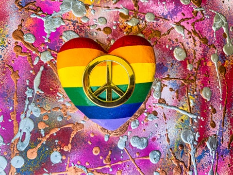 A heart-shaped object painted with the rainbow colors of the LGBTQ+ pride flag, featuring a prominent gold peace symbol in the center. The background is a vibrant and chaotic mix of bright pinks, purples, and oranges splattered with metallic silver and gold textures.