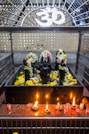 A shrine featuring black stone deities decorated with garlands of flowers is placed behind a metal enclosure. The area is adorned with yellow and white flowers, and there are several small oil lamps on a red granite surface in front of the deities. Above the shrine, there is a white Om symbol.