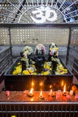 A shrine featuring black stone deities decorated with garlands of flowers is placed behind a metal enclosure. The area is adorned with yellow and white flowers, and there are several small oil lamps on a red granite surface in front of the deities. Above the shrine, there is a white Om symbol.