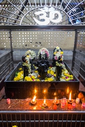 A shrine featuring black stone deities decorated with garlands of flowers is placed behind a metal enclosure. The area is adorned with yellow and white flowers, and there are several small oil lamps on a red granite surface in front of the deities. Above the shrine, there is a white Om symbol.