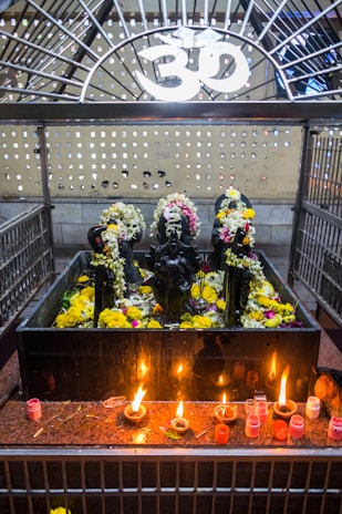 A shrine featuring black stone deities decorated with garlands of flowers is placed behind a metal enclosure. The area is adorned with yellow and white flowers, and there are several small oil lamps on a red granite surface in front of the deities. Above the shrine, there is a white Om symbol.