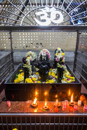 A shrine featuring black stone deities decorated with garlands of flowers is placed behind a metal enclosure. The area is adorned with yellow and white flowers, and there are several small oil lamps on a red granite surface in front of the deities. Above the shrine, there is a white Om symbol.