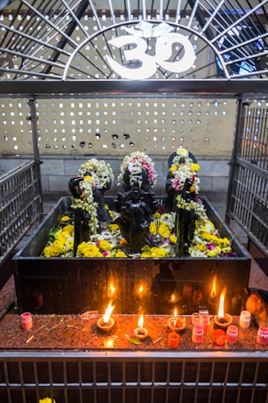 A shrine featuring black stone deities decorated with garlands of flowers is placed behind a metal enclosure. The area is adorned with yellow and white flowers, and there are several small oil lamps on a red granite surface in front of the deities. Above the shrine, there is a white Om symbol.
