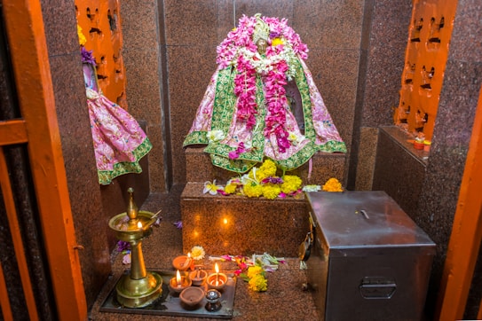 A sacred space with a statue adorned in vibrant pink and green fabric embellished with floral patterns. The statue is further decorated with garlands of fresh flowers. The setting includes a brass lamp with lit candles, an arrangement of various colored flowers, and a shiny metal donation box. The walls are lined with carved niches that hold small items and more flowers.