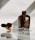 A brown bottle of repair essence hydrating moisturizer is placed upright on a reflective surface. The glass dropper, partially filled with liquid, lies next to the bottle, with reflections visible on the surface. The bottle bears a label with text, indicating it is a skincare product.