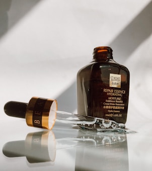 A brown bottle of repair essence hydrating moisturizer is placed upright on a reflective surface. The glass dropper, partially filled with liquid, lies next to the bottle, with reflections visible on the surface. The bottle bears a label with text, indicating it is a skincare product.