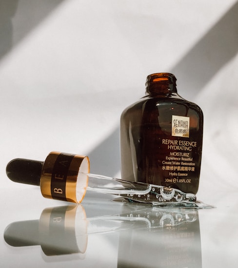 A brown bottle of repair essence hydrating moisturizer is placed upright on a reflective surface. The glass dropper, partially filled with liquid, lies next to the bottle, with reflections visible on the surface. The bottle bears a label with text, indicating it is a skincare product.