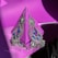 A pair of ornate, silver earrings with intricate filigree design and blue gemstone accents are displayed against a vibrant purple background. The earrings have an oval pattern, each adorned with several blue jewels, and they hang from a stand that casts some shadow. The silver material and the gemstones reflect light, creating a bright and eye-catching contrast against the purple backdrop.