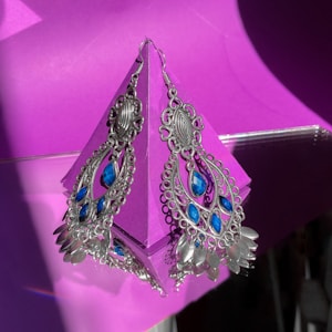 A pair of ornate, silver earrings with intricate filigree design and blue gemstone accents are displayed against a vibrant purple background. The earrings have an oval pattern, each adorned with several blue jewels, and they hang from a stand that casts some shadow. The silver material and the gemstones reflect light, creating a bright and eye-catching contrast against the purple backdrop.