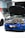 A blue sports car with its hood open reveals a complex engine inside a garage. Two other cars are partially visible, with one car being white and the other grey. The environment has a garage door slightly open and various tools and equipment typical of an auto repair or tuning shop.