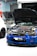 A blue sports car with its hood open reveals a complex engine inside a garage. Two other cars are partially visible, with one car being white and the other grey. The environment has a garage door slightly open and various tools and equipment typical of an auto repair or tuning shop.