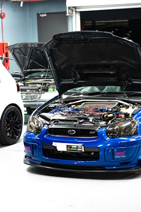 A blue sports car with its hood open reveals a complex engine inside a garage. Two other cars are partially visible, with one car being white and the other grey. The environment has a garage door slightly open and various tools and equipment typical of an auto repair or tuning shop.