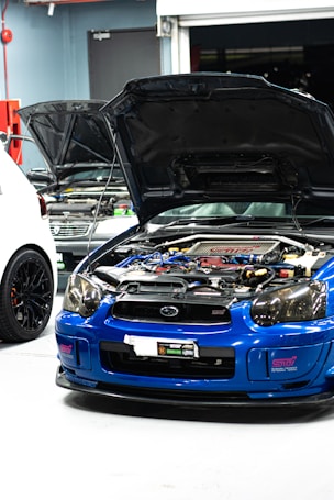 A blue sports car with its hood open reveals a complex engine inside a garage. Two other cars are partially visible, with one car being white and the other grey. The environment has a garage door slightly open and various tools and equipment typical of an auto repair or tuning shop.