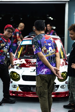 Several people are gathered around a car with vibrant decals and a colorful hood. They appear to be in a garage setting. The individuals are wearing bright, patterned shirts with cartoonish designs and logos.