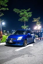 A blue compact car is parked on the side of a dimly lit street at night. The car has a license plate reading 'BKP 2059' and is surrounded by a few trees illuminated by streetlights. Several people are standing nearby in the background.
