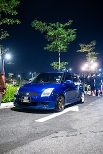 A blue compact car is parked on the side of a dimly lit street at night. The car has a license plate reading 'BKP 2059' and is surrounded by a few trees illuminated by streetlights. Several people are standing nearby in the background.