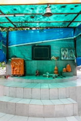 A small shrine is set on a multi-tiered white tiled platform. It features religious symbols, including an orange and black sacred stone or idol, a bell hanging from the roof, and offerings such as an orange vase and other small items. There is a framed image of a deity on the right side of the shrine. The area is sheltered by a turquoise tarpaulin roof that casts a greenish hue over the scene.