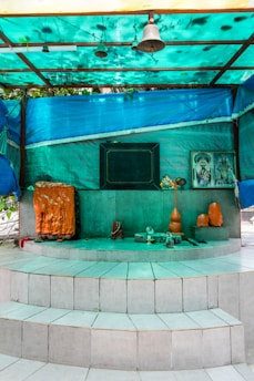A small shrine is set on a multi-tiered white tiled platform. It features religious symbols, including an orange and black sacred stone or idol, a bell hanging from the roof, and offerings such as an orange vase and other small items. There is a framed image of a deity on the right side of the shrine. The area is sheltered by a turquoise tarpaulin roof that casts a greenish hue over the scene.
