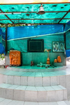 A small shrine is set on a multi-tiered white tiled platform. It features religious symbols, including an orange and black sacred stone or idol, a bell hanging from the roof, and offerings such as an orange vase and other small items. There is a framed image of a deity on the right side of the shrine. The area is sheltered by a turquoise tarpaulin roof that casts a greenish hue over the scene.