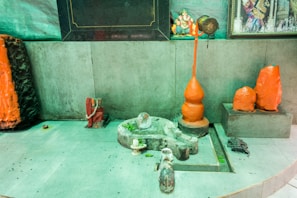 A shrine featuring religious statues and symbols, including a large orange trident and various stone carvings. The walls are lined with other religious icons and artifacts, with some being draped in fabric. The setting suggests a place of worship or meditation.