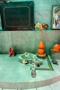 A small shrine area featuring a gray stone structure resembling a Lingam and Yoni, accompanied by small figurines and orange objects. A figurine of Ganesha sits to the right, alongside colorful religious images and statues. The setting has a spiritual and sacred ambiance.