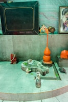 A small shrine area featuring a gray stone structure resembling a Lingam and Yoni, accompanied by small figurines and orange objects. A figurine of Ganesha sits to the right, alongside colorful religious images and statues. The setting has a spiritual and sacred ambiance.
