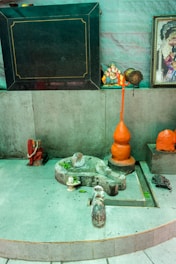 A small shrine area featuring a gray stone structure resembling a Lingam and Yoni, accompanied by small figurines and orange objects. A figurine of Ganesha sits to the right, alongside colorful religious images and statues. The setting has a spiritual and sacred ambiance.