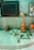 A small shrine area featuring a gray stone structure resembling a Lingam and Yoni, accompanied by small figurines and orange objects. A figurine of Ganesha sits to the right, alongside colorful religious images and statues. The setting has a spiritual and sacred ambiance.