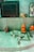 A small shrine area featuring a gray stone structure resembling a Lingam and Yoni, accompanied by small figurines and orange objects. A figurine of Ganesha sits to the right, alongside colorful religious images and statues. The setting has a spiritual and sacred ambiance.