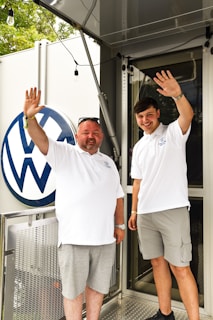 Two men in white polo shirts stand together waving and smiling, with a Volkswagen logo visible in the background. One of the men is older and wearing sunglasses, while the other is younger and wearing shorts.