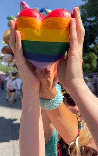 Hands are holding a heart-shaped object painted with rainbow colors. People in the background appear to be at an outdoor event under a clear blue sky.