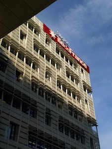 A modern building with a grid-like facade and numerous windows is prominently featured. The structure is bathed in sunlight, which creates shadows on its exterior. A large sign displaying 'Région Île-de-France' is mounted on the building, with a red star symbol. The sky in the background is clear and blue.