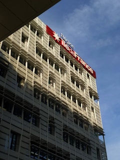 A modern building with a grid-like facade and numerous windows is prominently featured. The structure is bathed in sunlight, which creates shadows on its exterior. A large sign displaying 'Région Île-de-France' is mounted on the building, with a red star symbol. The sky in the background is clear and blue.