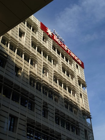 A modern building with a grid-like facade and numerous windows is prominently featured. The structure is bathed in sunlight, which creates shadows on its exterior. A large sign displaying 'Région Île-de-France' is mounted on the building, with a red star symbol. The sky in the background is clear and blue.