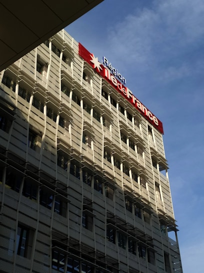 A modern building with a grid-like facade and numerous windows is prominently featured. The structure is bathed in sunlight, which creates shadows on its exterior. A large sign displaying 'Région Île-de-France' is mounted on the building, with a red star symbol. The sky in the background is clear and blue.