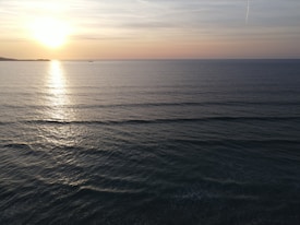 A serene ocean view at sunset with gentle waves reflecting the orange and purple hues of the sky. The sun is near the horizon, casting a warm glow on the water. A small silhouette of a boat is visible in the distance.
