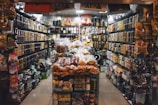 A small, crowded shop filled with a variety of packaged food items. Shelves are stacked with nuts, snacks, and other edible goods in jars and plastic bags. The shop has a narrow aisle leading to the back where more products are visible, including colorful bags and boxes.