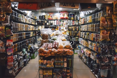 A small, crowded shop filled with a variety of packaged food items. Shelves are stacked with nuts, snacks, and other edible goods in jars and plastic bags. The shop has a narrow aisle leading to the back where more products are visible, including colorful bags and boxes.