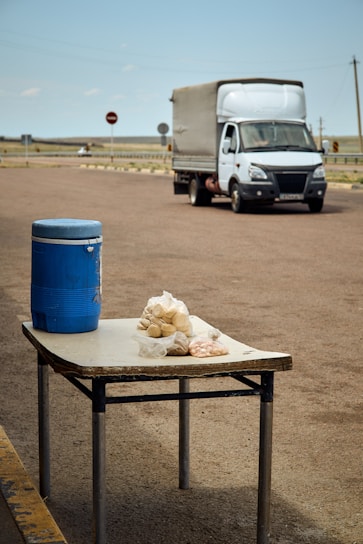 A roadside stall features a blue insulated jug and bags of food on a worn table, with a white delivery truck passing by in the background. The road is deserted, surrounded by flat terrain with sparse markers and signs under a clear sky.