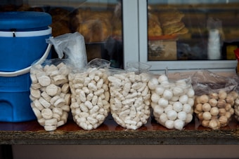Several bags filled with popcorn and various snacks are displayed on a counter. A blue cooler with a white lid and handle is positioned to the left. In the background, there are more snacks and plastic cups visible through a window.