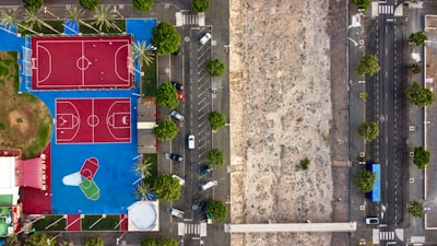 Aerial view of a sports complex with red courts on a bright blue surface, located next to a large open area and a road lined with parked cars and green trees.