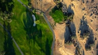 An aerial view of a golf course with lush green grass, sand bunkers, and long shadows cast by tall palm trees. The vibrant green contrasts with the surrounding sandy and rocky terrain typical of a desert landscape.