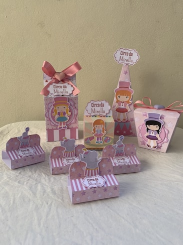 A collection of decorative party favor boxes with a circus theme featuring cartoon characters. They are adorned with images of girls in circus attire and small elephants wearing hats. The boxes are primarily pink with accents of white and light purple, some featuring star patterns. A large pink bow is prominent on one of the boxes.