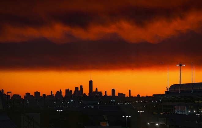 A dramatic skyline with a vivid orange and red sunset. The city silhouette includes a variety of skyscrapers, while dark clouds loom overhead. Tiny airplanes can be seen flying above the cityscape, adding to the scene's sense of scale.