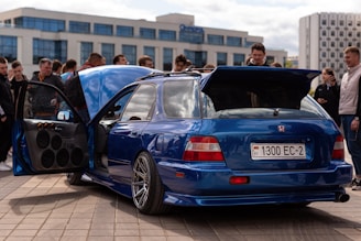 A modified blue sports car with custom wheels and an open door displaying a powerful sound system. Several people stand around, seemingly admiring the vehicle. The car is parked on a paved area in an urban setting with modern buildings in the background.